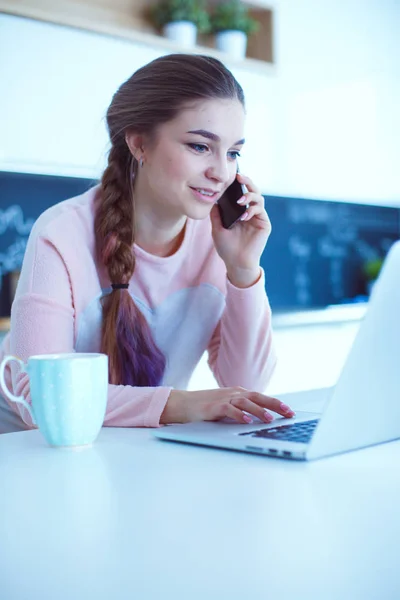 Junge Frau sitzt mit Laptop am Küchentisch und telefoniert. — Stockfoto