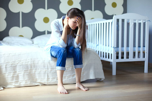 Young tired woman sitting on the bed near childrens cot. Young mom.