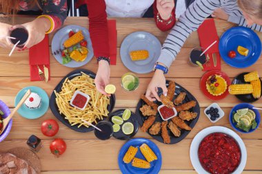 Ahşap masada otururken birlikte yemek yiyen bir grup insanın üst düzey görüntüsü. Masada yemek var. İnsanlar fast food yiyor..