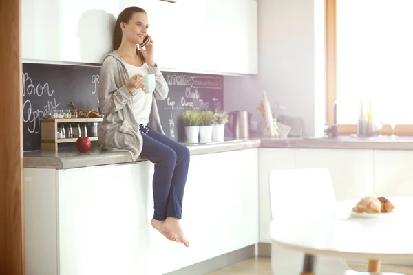 Mulher usando telefone celular sentado na cozinha moderna . — Fotografia de Stock