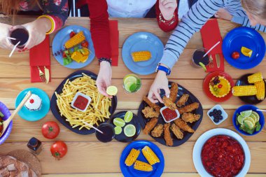 Ahşap masada otururken birlikte yemek yiyen bir grup insanın üst düzey görüntüsü. Masada yemek var. İnsanlar fast food yiyor..