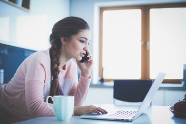 Genç kadın bir dizüstü bilgisayar kullanarak ve cep telefonuyla konuşurken mutfak masasında oturur.