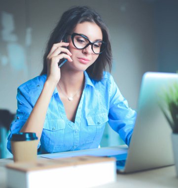 Güzel genç iş kadını ofis masasında oturuyor ve cep telefonuyla konuşuyor.