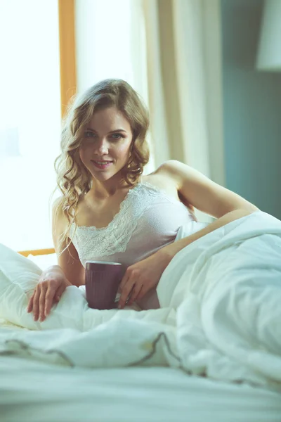Νεαρή γυναίκα πίνοντας καφέ ή τσάι ξαπλωμένοι στο κρεβάτι. — Φωτογραφία Αρχείου