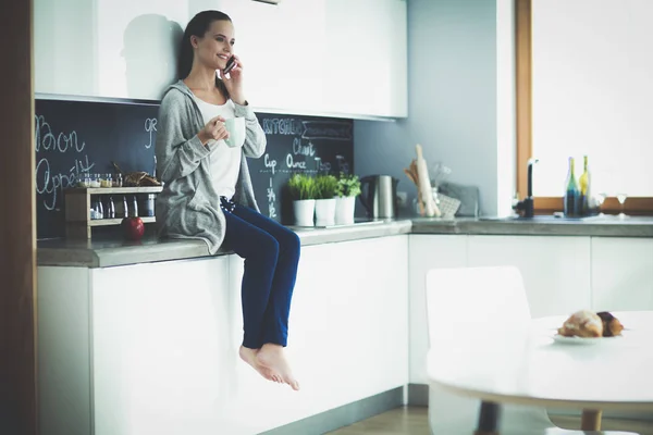 Mujer usando el teléfono móvil sentado en la cocina moderna . — Foto de Stock