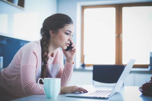 Une jeune femme s'assoit à la table de la cuisine en utilisant un ordinateur portable et en parlant sur un téléphone portable . — Photo