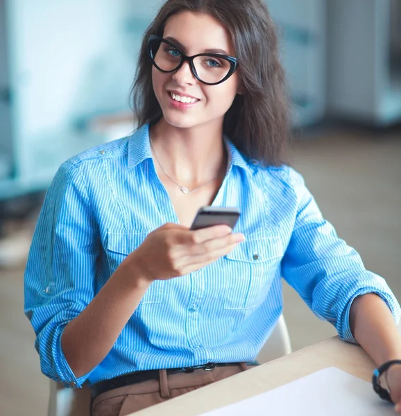 Gyönyörű fiatal üzletasszony ül irodai íróasztal és a kezében mobiltelefon — Stock Fotó