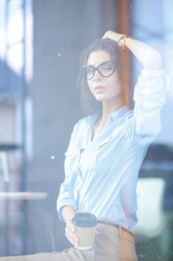 Ofis masasında dizüstü bilgisayarıyla oturan genç bir kadın. Pencereden bakıyor. Genç kadın.