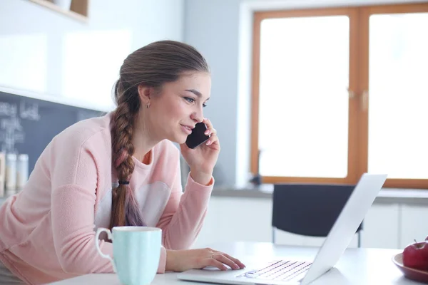 Une jeune femme s'assoit à la table de la cuisine en utilisant un ordinateur portable et en parlant sur un téléphone portable . — Photo