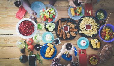 Ahşap masada otururken birlikte yemek yiyen bir grup insanın üst düzey görüntüsü. Masada yemek var. İnsanlar fast food yiyor..