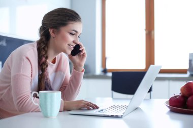 Genç kadın bir dizüstü bilgisayar kullanarak ve cep telefonuyla konuşurken mutfak masasında oturur.