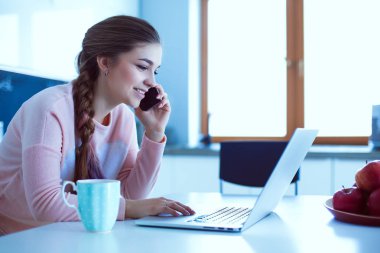 Genç kadın bir dizüstü bilgisayar kullanarak ve cep telefonuyla konuşurken mutfak masasında oturur.