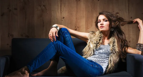 优雅的女人坐在黑色的沙发上，穿着一件蓝色牛仔裤的肖像和毛皮背心 — 图库照片