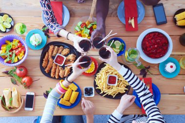 Ahşap masada otururken birlikte yemek yiyen bir grup insanın üst düzey görüntüsü. Masada yemek var. İnsanlar fast food yiyor..