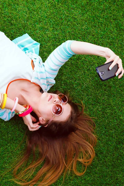 Mooie jonge vrouw die selfie maakt door haar telefoon terwijl ze in groen gras ligt. mooie jonge vrouw maken selfie — Stockfoto