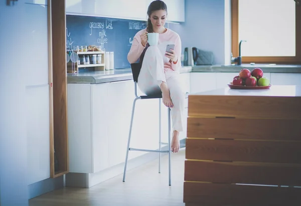 Mujer usando el teléfono móvil sentado en la cocina moderna . — Foto de Stock