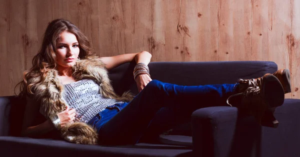 Portrait of elegant woman sitting on black sofa wearing a blue jeans and fur vest — Stock Photo, Image