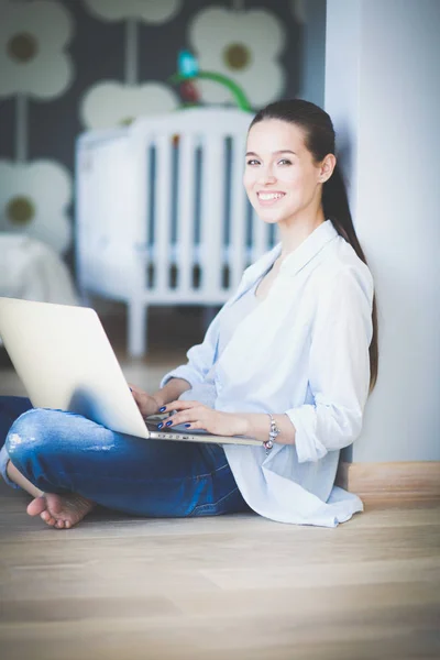 Ung kvinna som sitter på golvet nära barnens babysäng med laptop. Ung mamma — Stockfoto