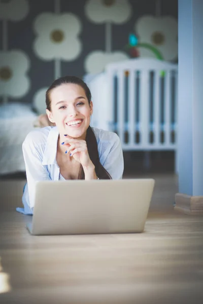 Ung kvinna som sitter på golvet nära barnens babysäng med laptop. Ung mamma — Stockfoto