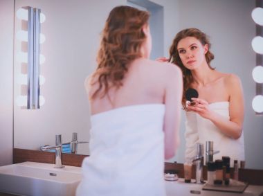 Genç bir kadın aynaya bakıyor ve makyaj yapıyor..