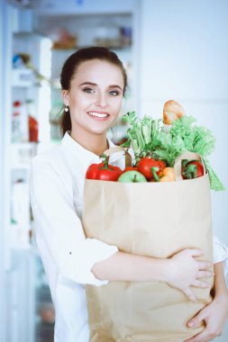Sebzeli alışveriş torbası tutan genç bir kadın. Mutfakta duruyor. Mutfakta kameraya bakan bir kadın var.