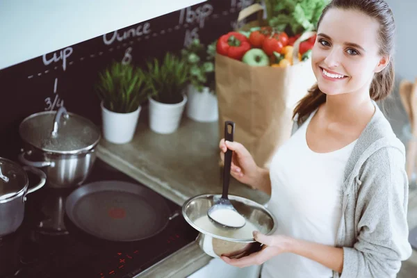 Νεαρή γυναίκα ετοιμάζει τηγανίτες στην κουζίνα, ενώ στέκεται κοντά στο τραπέζι. Γυναίκα στην κουζίνα. Μαγειρική στην κουζίνα. — Φωτογραφία Αρχείου