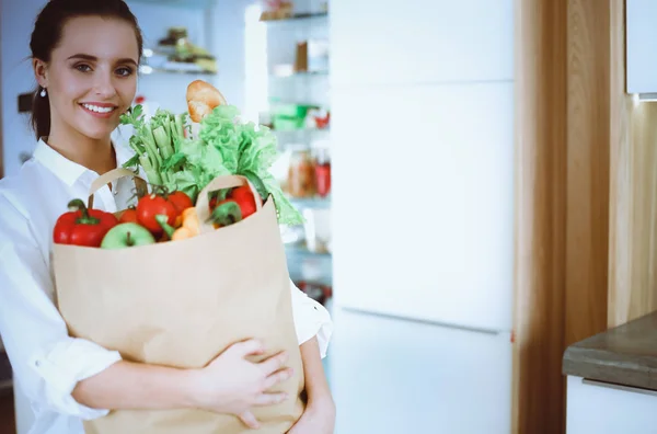 Jeune femme tenant sac d'épicerie avec des légumes. Debout dans la cuisine. Femme dans la cuisine regardant la caméra — Photo