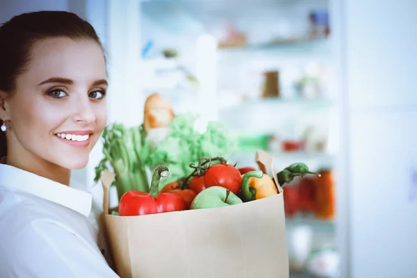 Jeune femme tenant sac d'épicerie avec des légumes. Debout dans la cuisine. Femme dans la cuisine regardant la caméra — Photo