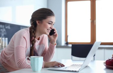 Genç kadın bir dizüstü bilgisayar kullanarak ve cep telefonuyla konuşurken mutfak masasında oturur.