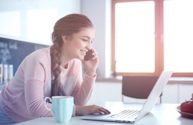 Genç kadın bir dizüstü bilgisayar kullanarak ve cep telefonuyla konuşurken mutfak masasında oturur.