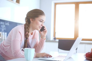 Genç kadın bir dizüstü bilgisayar kullanarak ve cep telefonuyla konuşurken mutfak masasında oturur.