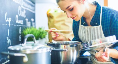 Genç bir kadın mutfağında yemek pişiriyor sobanın yanında duruyor.