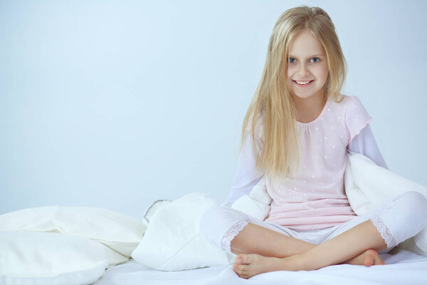 Little girl in the bedroom is sitting on the bed. Little girl is wearing a pajamas and sitting in bed