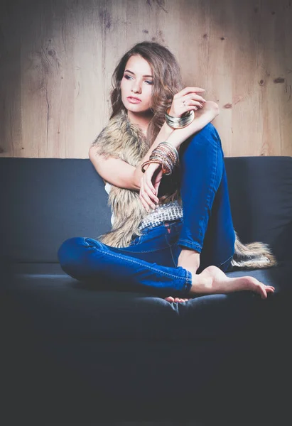 Portrait of elegant woman sitting on black sofa wearing a blue jeans and fur vest — Stock Photo, Image