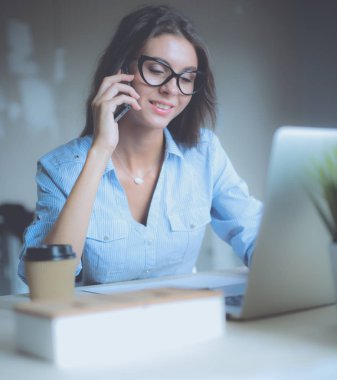 Güzel genç iş kadını ofis masasında oturuyor ve cep telefonuyla konuşuyor.