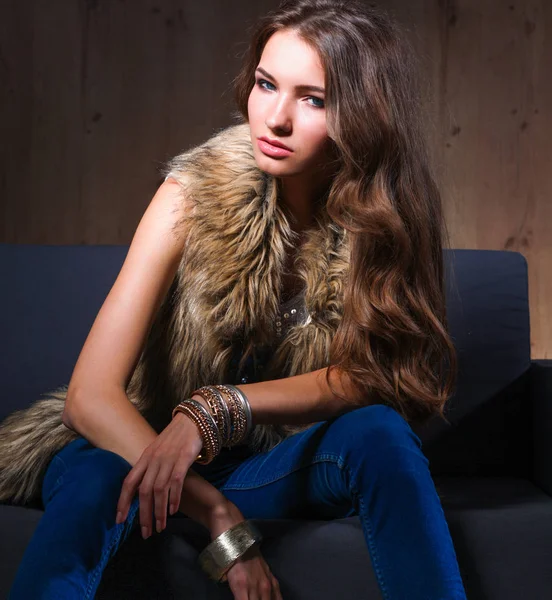 Portrait of elegant woman sitting on black sofa wearing a blue jeans and fur vest — Stock Photo, Image