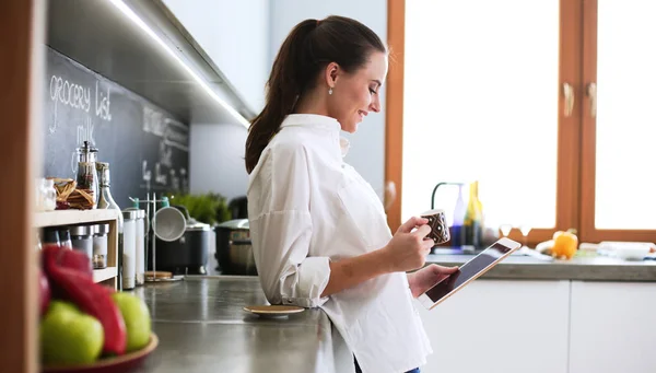 Mujer joven usando la tableta en la cocina en casa y beber café —  Fotos de Stock