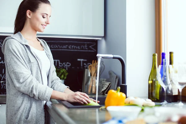 Schöne junge Frau wäscht Gemüse für Salat, während sie in der Küche steht. — Stockfoto