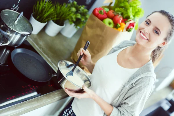 Jonge vrouw bereidt pannenkoeken in de keuken terwijl ze naast de tafel staat. Vrouw in de keuken. Koken in de keuken. — Stockfoto