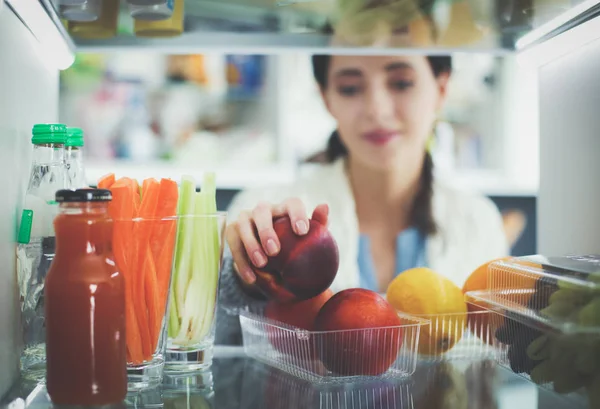 Портрет женщины, стоящей рядом с открытым холодильником, полным здоровой пищи, овощей и фруктов — стоковое фото