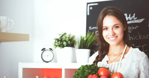 Mujer joven sonriente sosteniendo verduras de pie en la cocina —  Fotos de Stock