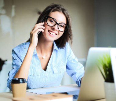 Güzel genç iş kadını ofis masasında oturuyor ve cep telefonuyla konuşuyor.