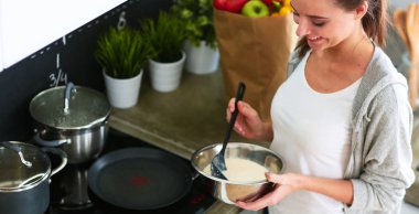 Genç bir kadın mutfakta krep pişiriyor sobanın yanında duruyor.