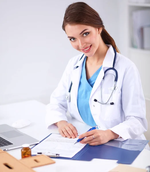 Vacker ung leende kvinnlig läkare sitter vid skrivbordet och skriver. — Stockfoto