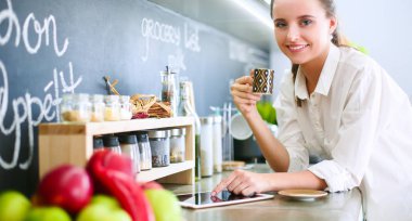 Genç bir kadın mutfakta tablet kullanıyor ve kahve içiyor.