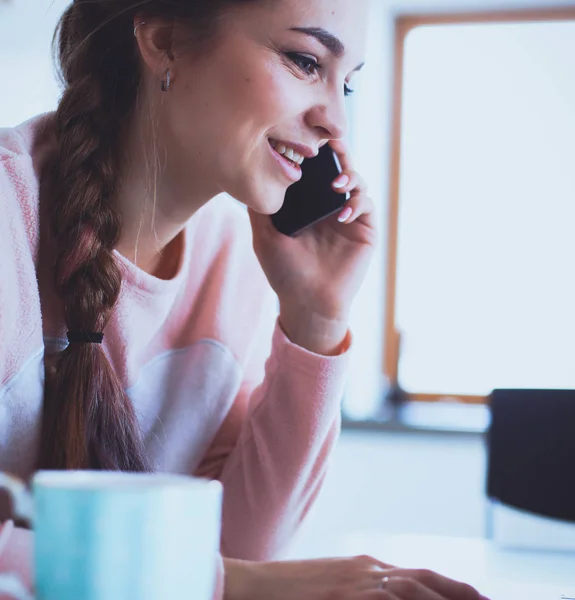 Une jeune femme s'assoit à la table de la cuisine en utilisant un ordinateur portable et en parlant sur un téléphone portable — Photo