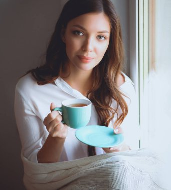 Evde oturan genç bir kadın oturma odasında oturmuş kitap okuyor ve çay ya da kahve içiyor.