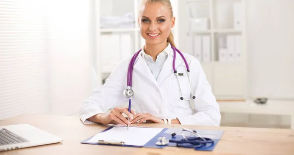 Bonito jovem sorridente médico feminino sentado na mesa e escrevendo. — Fotografia de Stock