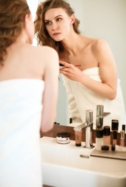 Genç bir kadın aynaya bakıyor ve makyaj yapıyor..