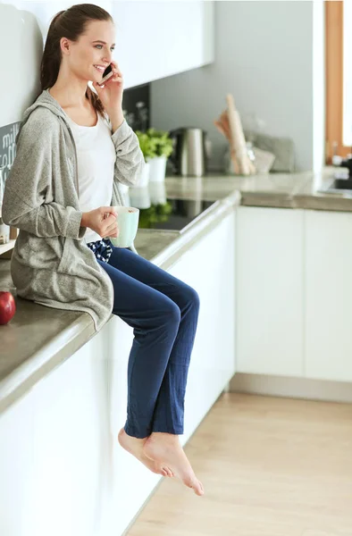 Mujer usando el teléfono móvil sentado en la cocina moderna . — Foto de Stock
